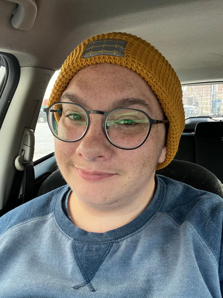 a selfie of the poster, a trans man with freckles and a septum piercing, wearing a yellow beanie and a pair of new black rimmed spectacles while smiling at the camera