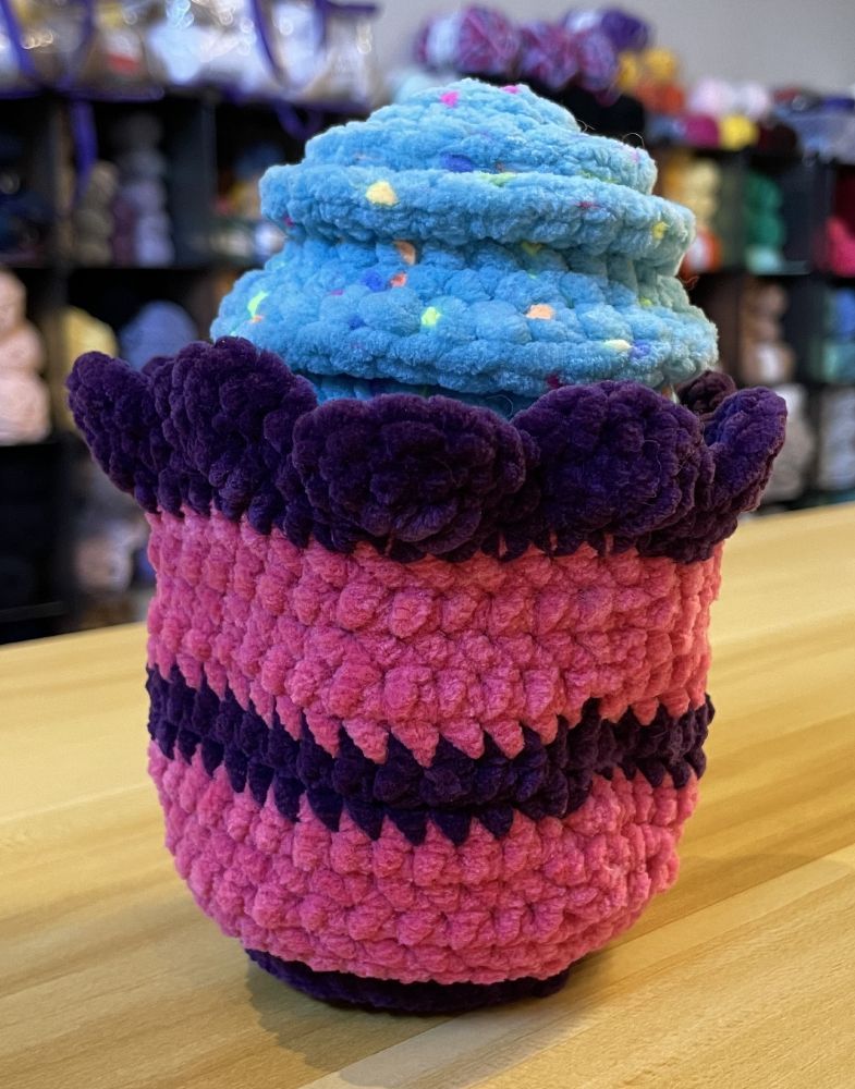 Crochet plushie in the form of a cupcake with a pink and dark purple wrapped and light blue frosting with sprinkles.