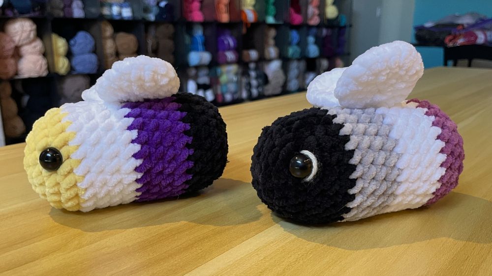 Side view of 2 small crochet plushie bees. The one on the left is made in the colors of the Nonbinary Pride flag and the one on the right is made in the colors of the Asexual Pride flag.