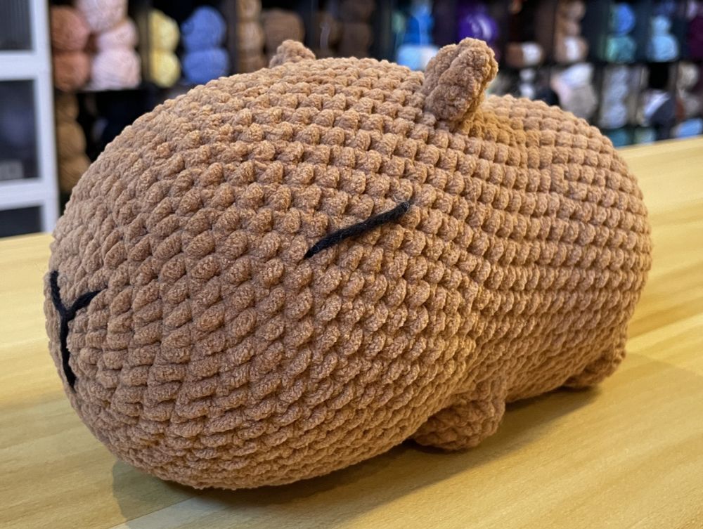 Partial profile view of a crochet plushie capybara made with light brown yarn with black embroidered nose and closed/sleeping eyes.