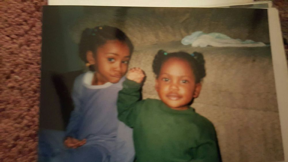 one year old nya is pictured in a green longsleeve crew neck, with one hand next to her face with her fingers curled in. her hair is (again) in puppy ponytails, styled as two twostrand twists that stop above her ear. she is looking into the camera.