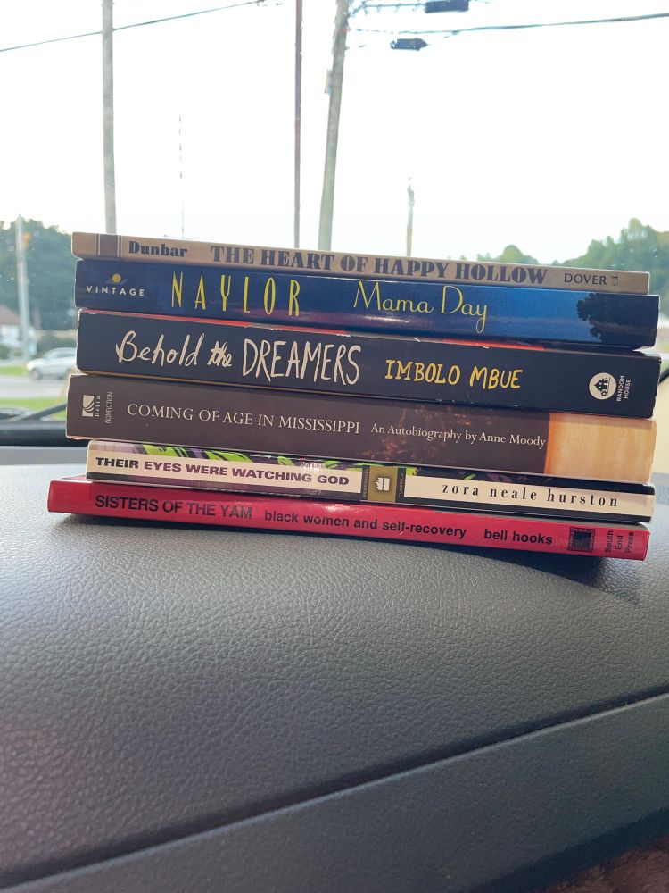 a stack of six books sitting atop a grey truck dashboard, spines facing the camera. from top to bottom:
The Heart of Happy Hollow by Paul Dunbar;
Naylor by Mama Day; Behold the Dreamers by Imbolo Mbue; Coming of Age in Mississippi by Anne Moody; Their Eyes Were Watching God by Zora Neale Hurston; and Sisters of the Yam by Bell Hooks.