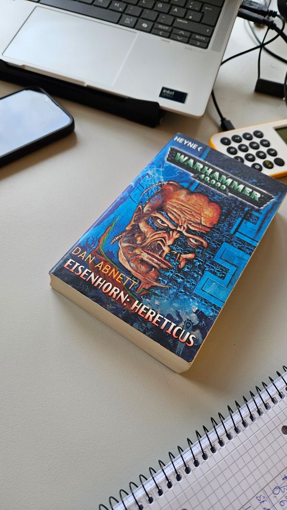 Alt-Text:
Ein Taschenbuch mit dem Titel „Warhammer 40,000 – Eisenhorn: Hereticus“ von Dan Abnett liegt auf einem Schreibtisch. Daneben befinden sich ein aufgeschlagenes kariertes Notizbuch, ein Smartphone, ein Laptop und ein Kartenlesegerät mit gelbem Rand.