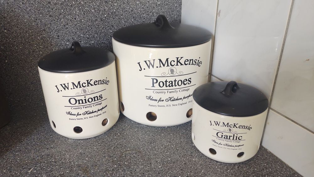 Witte J.W. McKensie potten met zwarte deksels: onions, potatoes en garlic uitgestald in een hoek van het aanrecht.