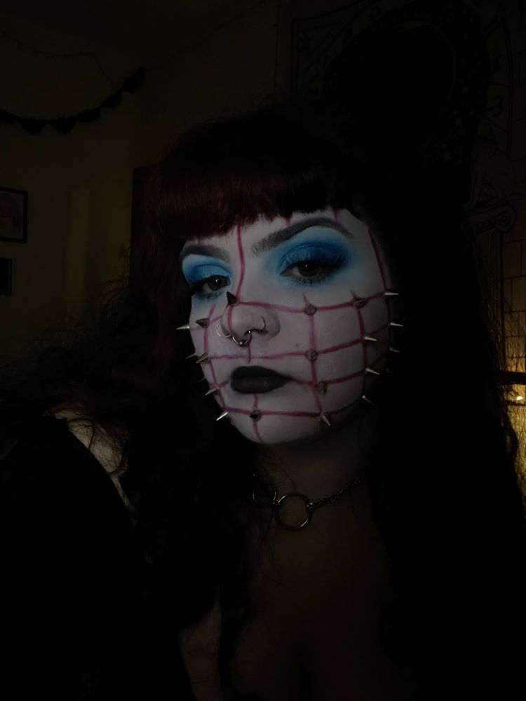 dark close up selfie of me as pinhead. I am wearing white face paint with blue eyeshadow, and a red grid on my face with spikes at each connecting point 