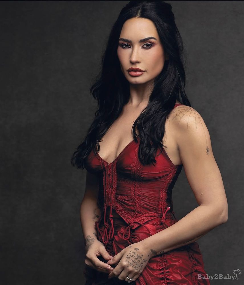 Photo of Demi Lovato in a red ruched top and skirt with their hair half up/half down. There’s a baby2baby watermark in the bottom right.