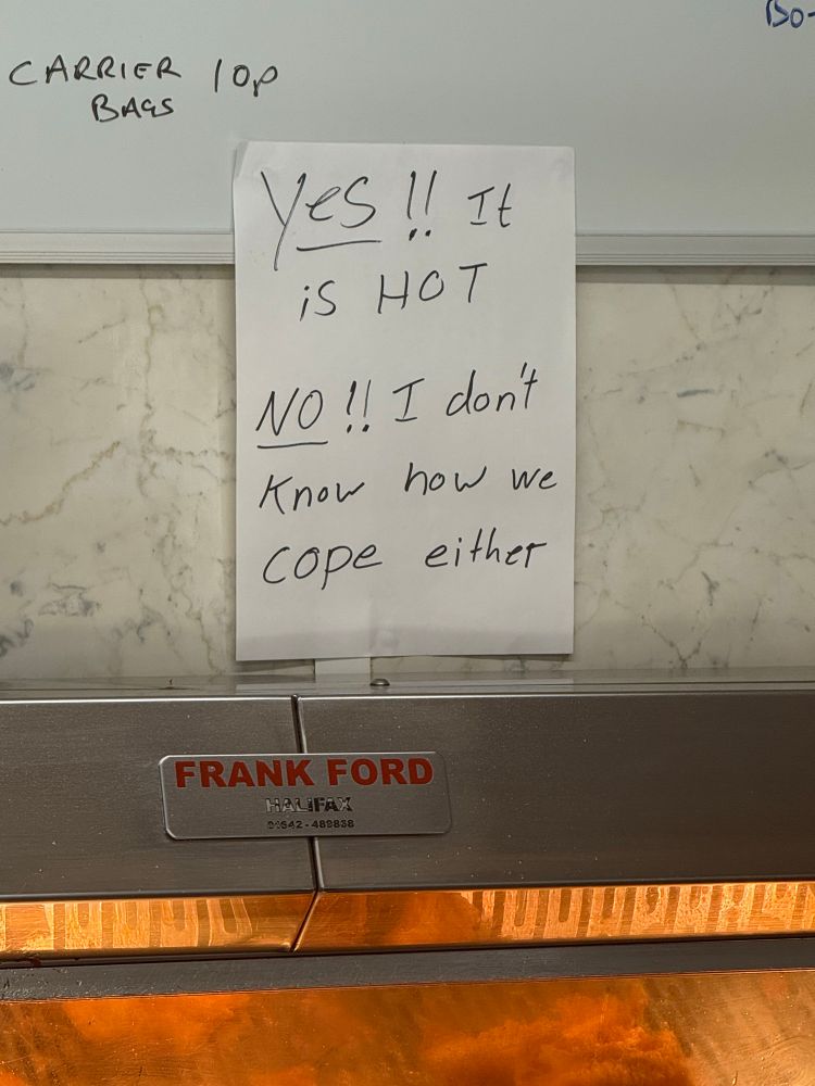 A sign pinned to the wall that reads “Yes! It is hot in here! And no, I don’t know how we cope either.” 