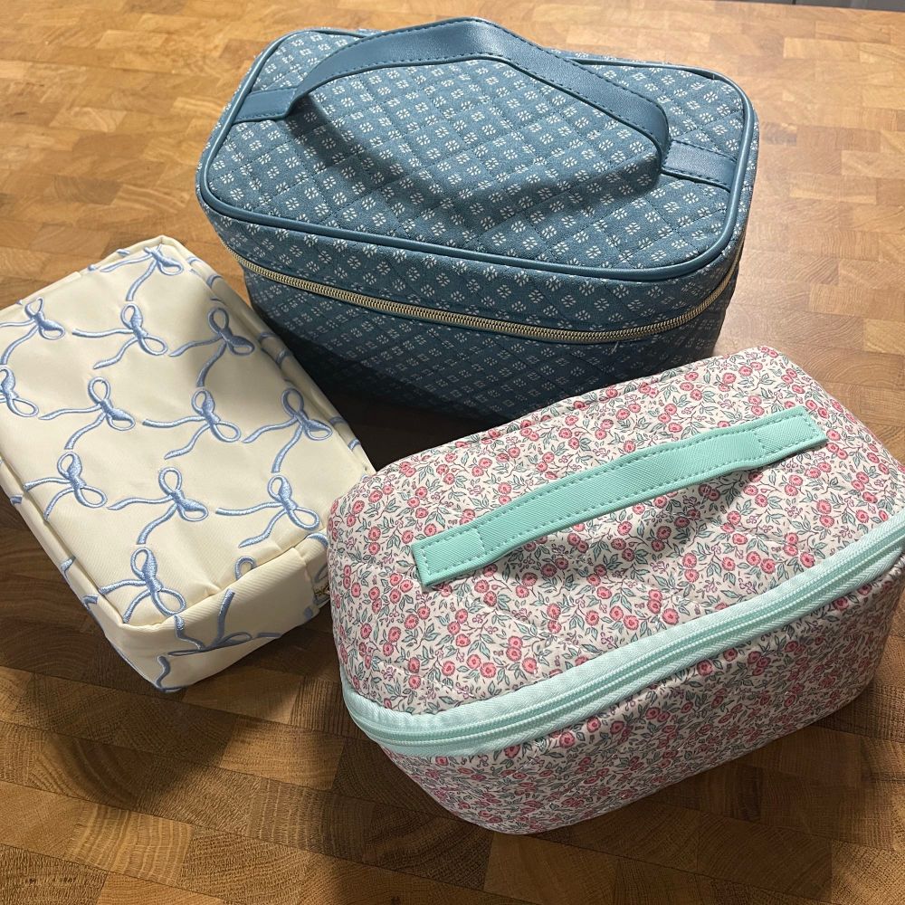Three toiletry bags sitting on a wood island table. Two have a quilted cotton finish, one with red flowers and a light blue trim and the other with a geometric dark blue and white diamond pattern. The third is white with blue embroidered bows. 