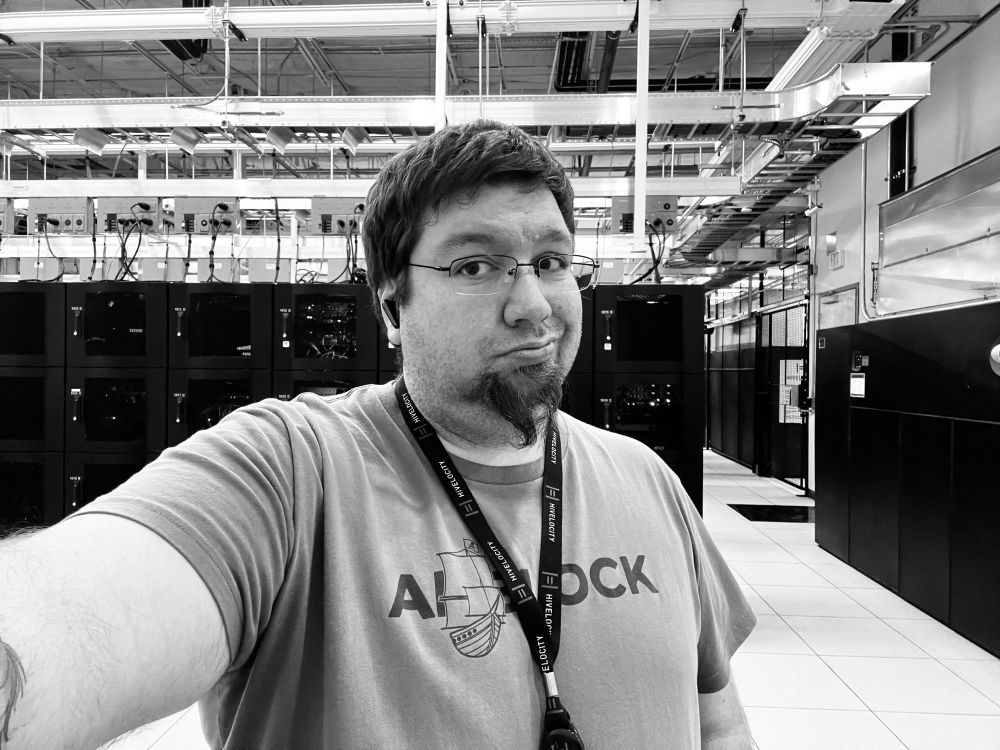 Black and white selfie of KJ with glasses and a goatee in a server room, making a slight frown. He is wearing a t-shirt with a boat graphic. The server room has rows of black server racks and the ceiling features a network of pipes and ventilation.