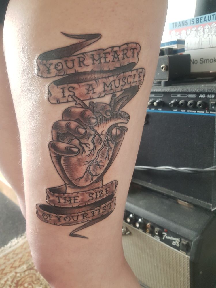 A side-on photo of someone's thigh with a fresh tattoo. It is in black and grey neo-traditional style and features a hand grasping an anatomical heart. A banner flowing above and below the hand reads "YOUR HEART IS A MUSCLE/THE SIZE OF YOUR FIST" in reference to a Ramshackle Glory song of the same name.