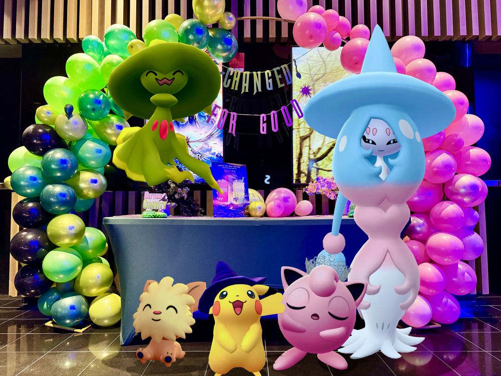 Witch Hat Pikachu, Jigglypuff wearing Glinda’s crown, Lillipup, Hatterene, and Shiny Green Mismagius smile for a photo in front of the “Wicked: For Good” at their local Regal Cinema theater. Behind them is an arch made of green and pink balloons and a banner that says “Changed for good”. Pokemon GO GOSnapshot AR Photography.