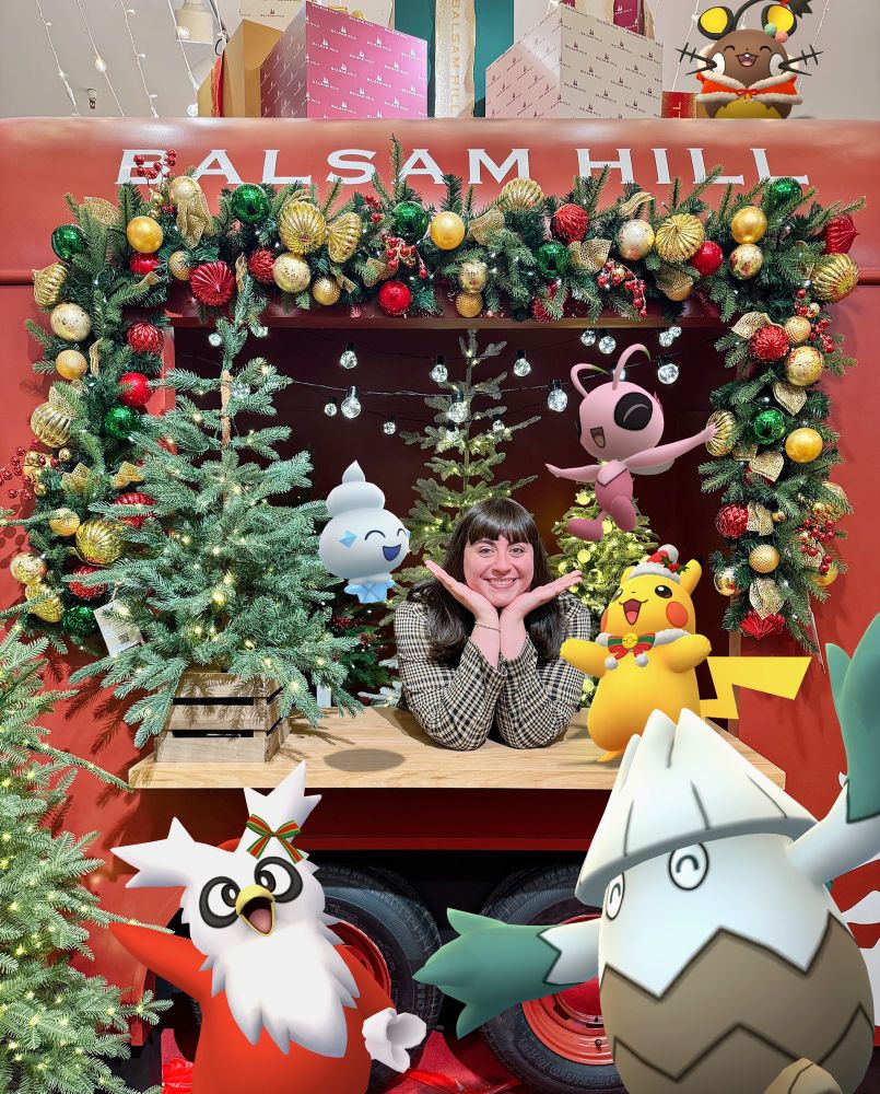 Julie on the 9th floor of Macy’s Department Store in New York City surrounded by decorations and garland. The location is decked out for the holiday season with Christmas merchandise and photo opportunities. She is joined by her Christmas team: Santa Hat Pikachu, Celebi, winter outfit Dedenne, Vanillite, Snover, and Delibird.