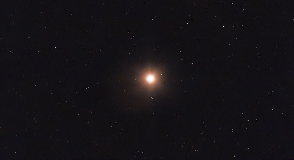 Mars, with a red/orange hue, looking like a bright shining star with smaller stars in the background.
