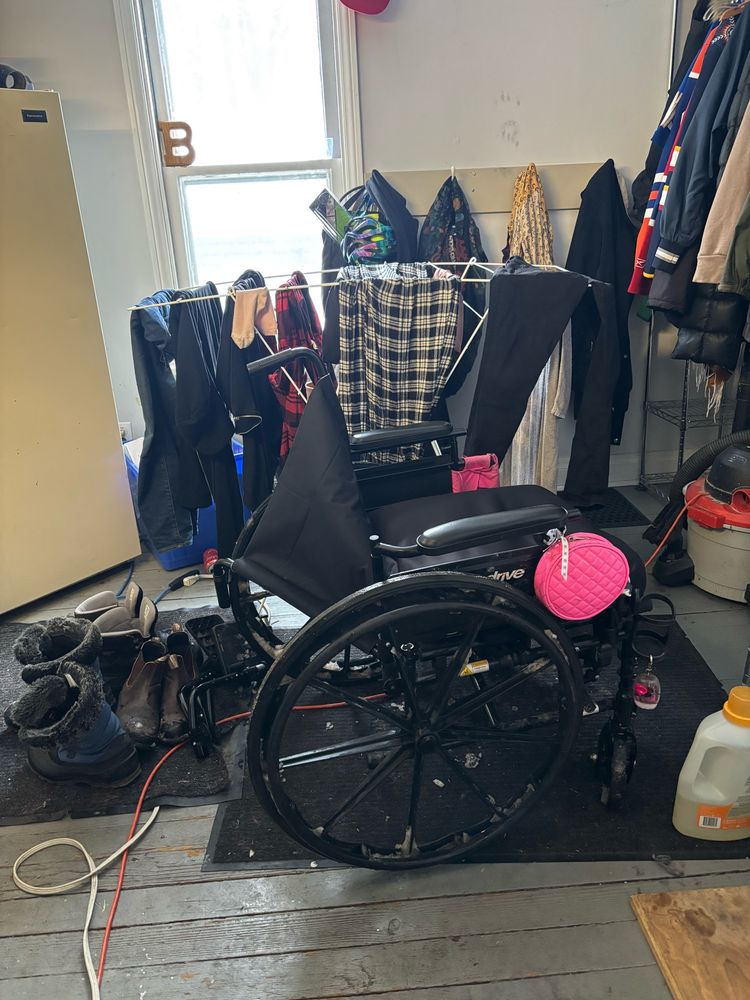 a side view of charis’ wheelchair cordelia. a shitty black manual chair with bright pink pockets attached. one of the metal poles supporting the back is recently broken on one side and the fabric back panel dangles  