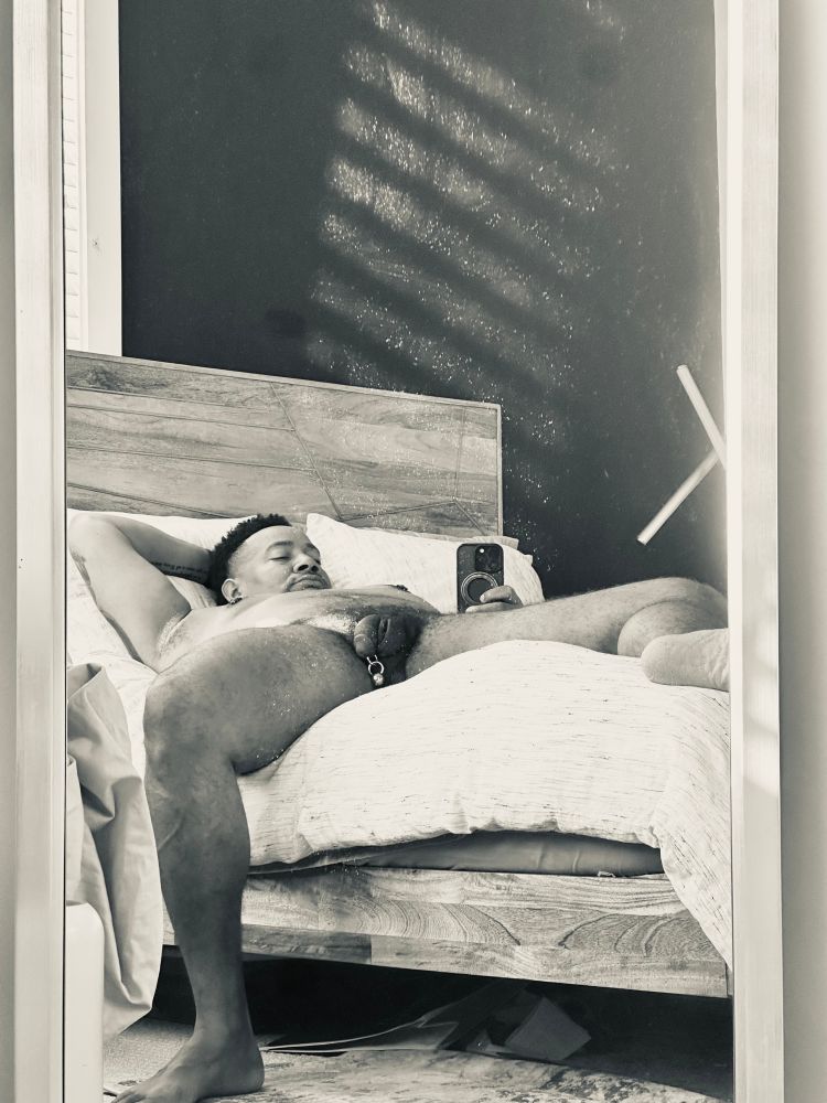 A silver tone photo of Dom laying down on his bed, holding his phone to take a picture of his reflection in the mirror. He’s got a large heavy ball hanging from his Prince Albert piercing 