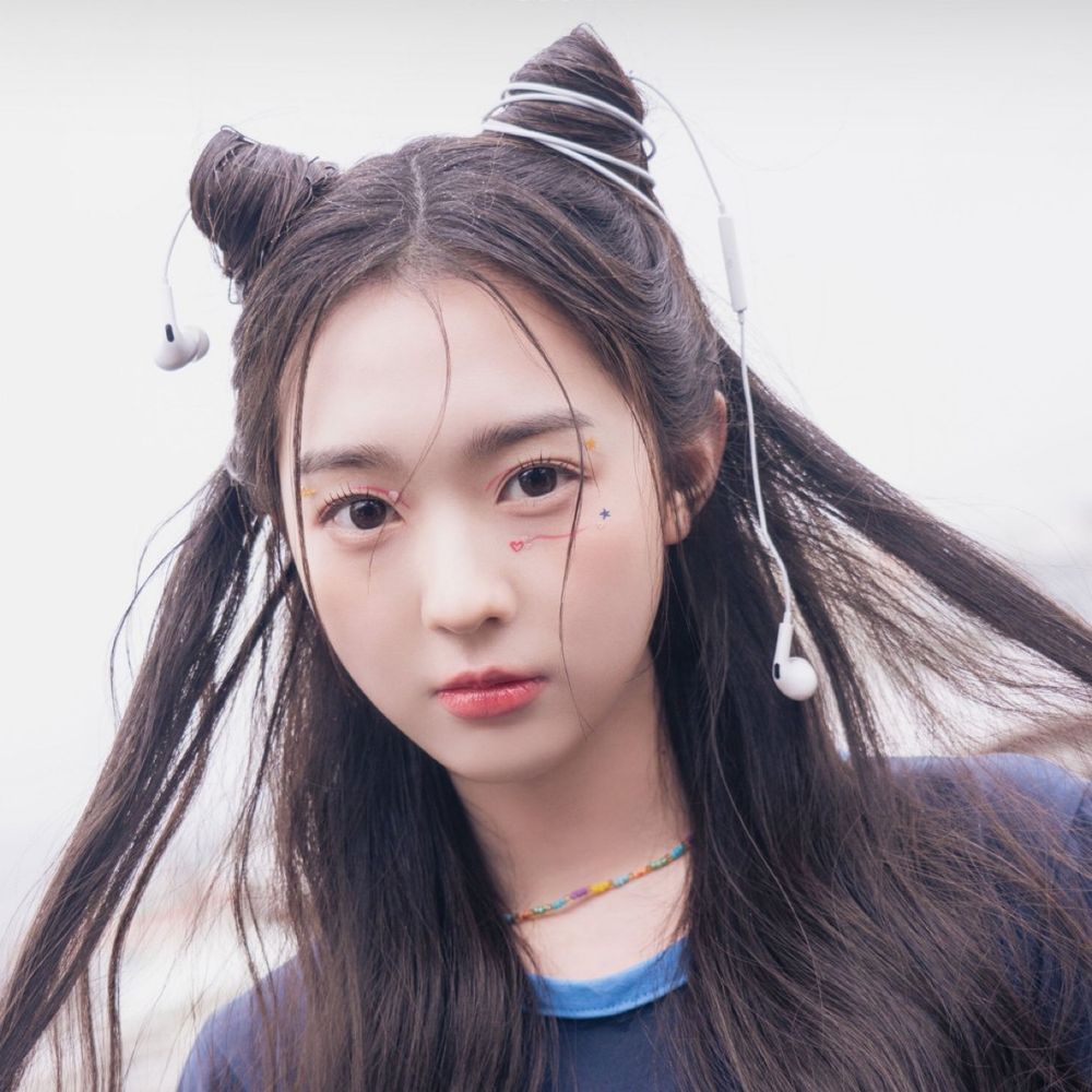 Soomin from tripleS with two buns in her hair that are shaped kind of like cat ears and have wired earbuds wrapped around them. She has little pink squiggles near her eyes and some tiny heart and star stickers.
