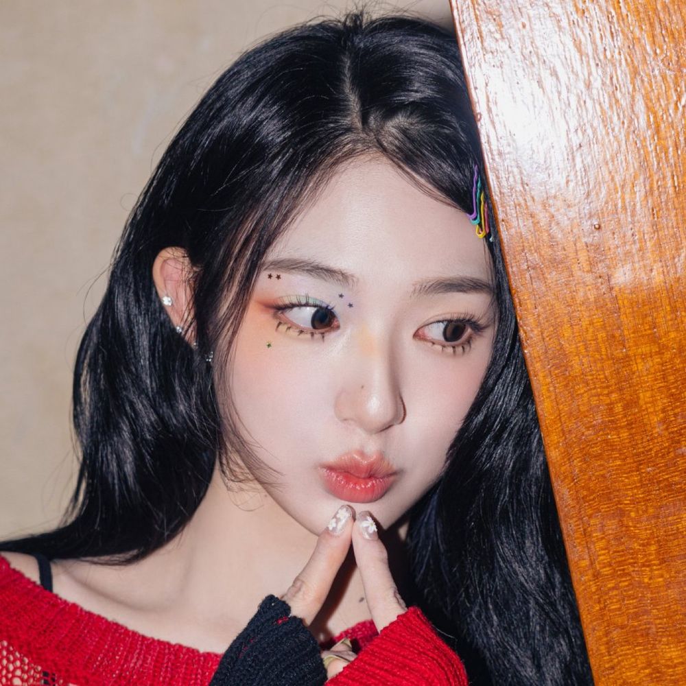 Xinyu from tripleS with rainbow eyeshadow and stars around her left eye and the other eye left mostly plain. There is a row of rainbow bobby pins on the right side of her hair though.
