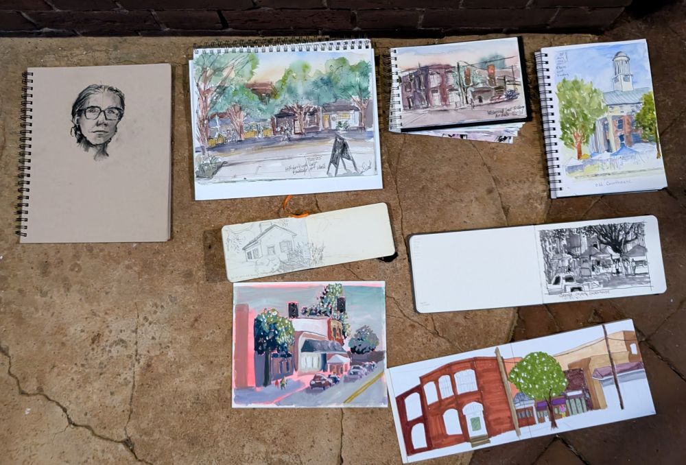 Multiple artist sketchbooks on the ground showing various scenes from a small town plus one portrait