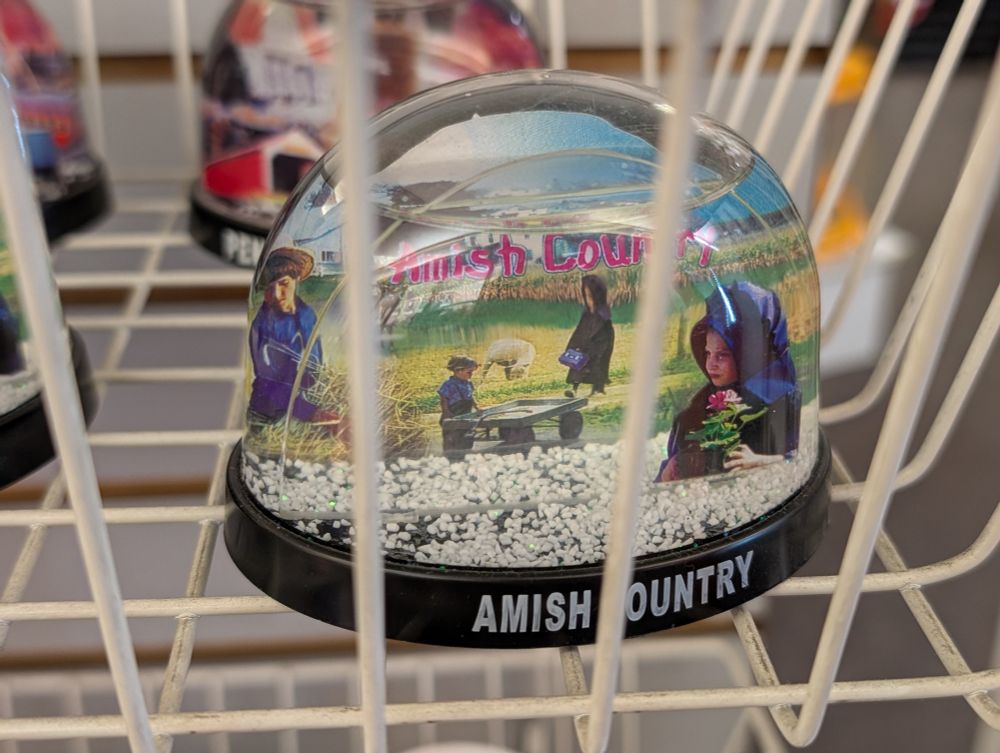 Snow globe labelled Amish Country. Inside are images of boys wearing straw hats and girls in bonnets in a farmland environment with corn and a sheep or goat. One boy gazes at a flat, empty wooden cart for unknown reasons. 