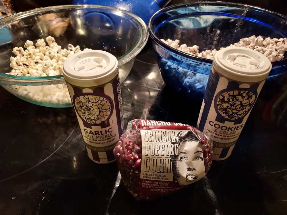 Two bowls of popcorn on a black glass-topped stove, one clear Pyrex and one blue Anchor Hocking.
In the middle are two small plastic canisters of popcorn seasonings: Garlic Dill Pickle and Cookies and Cream.
In the front is a half-empty bag of Rancho Gorgo Crimson Popping Corn.
10/10 for all!