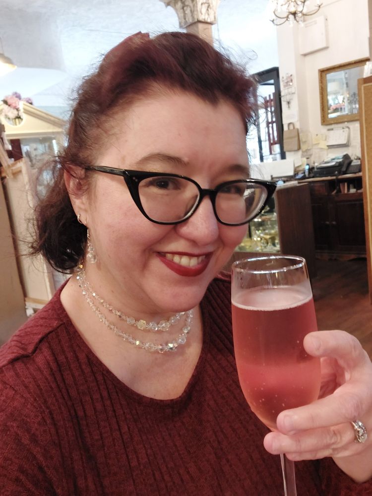 Me wearing black cat eye glasses and a burgundy rib knit dress and clear crystal double strand cocktail necklace. Holding a tall champagne flute glass of cranberry prosecco. Cheers!