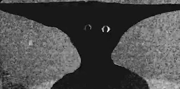 A weird little demon man from the Boisvert video 'Take Care'. He is entirely made of shadow with pale eyes staring at the viewer from an anvil-shaped head. 