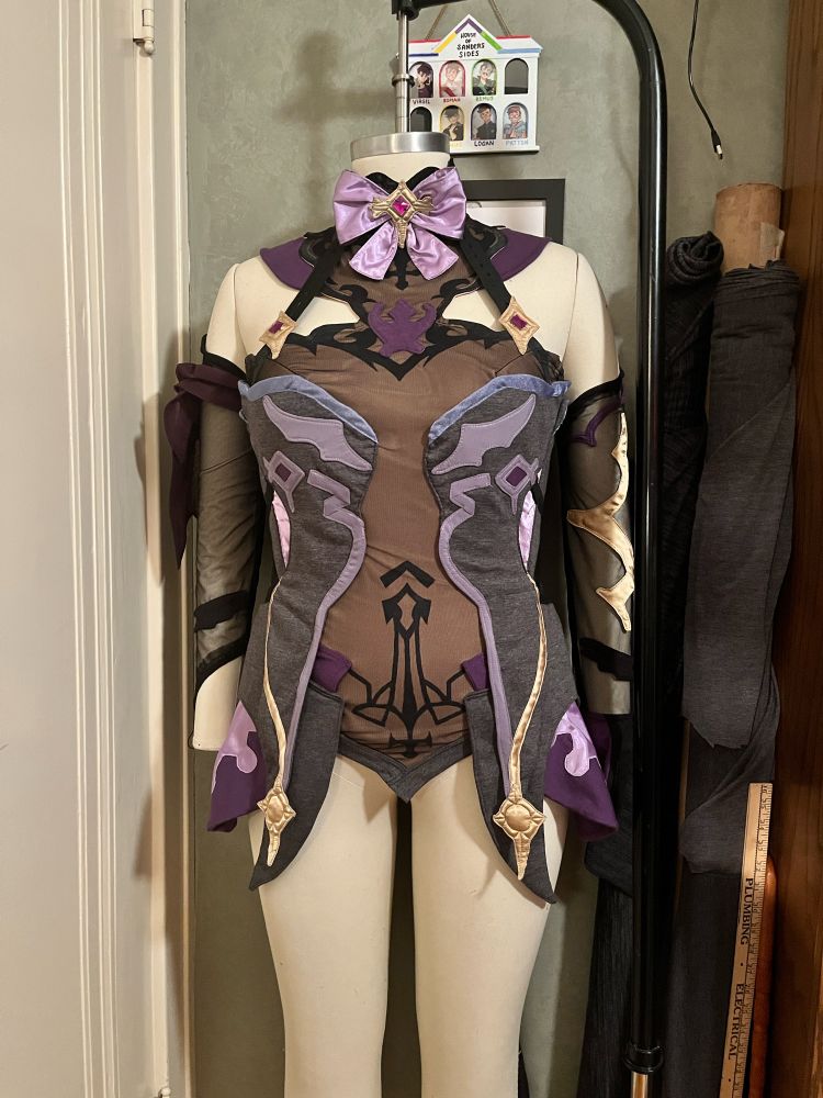 Cosplay commission for a close friend, completed in February 2024; Fischl from Genshin Impact.

Main pieces featured:
- a layered tunic with a pointed hem and high-low overskirt; ornate details made from lavender satin, dark purple jersey knit, heather grey jersey knit, purple-grey 4-way-stretch knit, tan ponte knit, black mesh, black fabric paint, periwinkle bias tape, purple sew-on gems, and gold satin. Back fastened with hooks & eyes and a zipper.

- a ruffled collar with large lavender bow; secured with ties for adjustability; made with lavender satin, black mesh, black jersey knit, and a gold satin star w/ a purple gem. The collar also has elastic straps that attach to the front and back necklines of the tunic for additional support.

- asymmetric gloves with curved top edge, both based in black mesh and features a small black wing and purple band. One glove also covers the hand using both black and purple jersey knit, and features a gold appliqué along the arm. The other glove is a fingerless-gauntlet style, and is layered under a purple-chiffon arm ruffle.

Other pieces, not pictured:
- black satin hair bows
- small black eyepatch
- leggings made from black jersey knit and black mesh; trim added along thighs using grey jersey knit