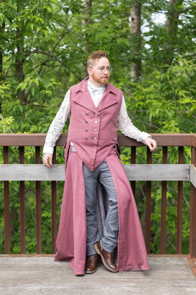 Wedding officiant, Zach Halliwell, wearing a sleeveless tailcoat and matching overskirt; commissioned winter/spring 2024

Both the tailcoat and overskirt are made from a heavyweight maroon linen and lined with a grey-floral printed cotton. The tailcoat features antique silver double-breasted buttons, a pointed front waistline, deep back pockets, an M-lapel, and an adjustable lace-up back (not pictured). The overskirt is a floor-length half circle with wrap-around waist ties, which are supported by belt loops and a buttonhole. The center back waistband has an inner tie to secure it to the back belt loop of the trousers worn underneath.