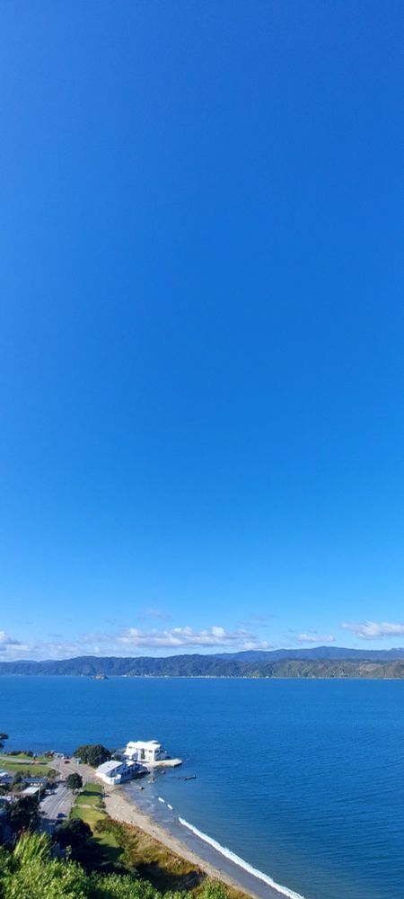 A view from the top of a hill overlooking the calm blue water of a bay and harbor, with lots of light, to dark blue sky. The only clouds are far off in the distant