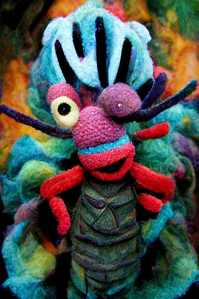 Vibrant handmade folk art creature with colorful felted wool body, featuring a whimsical face with one large eye, red mouth, and black hand-like appendages, set against a psychedelic tie-dye background