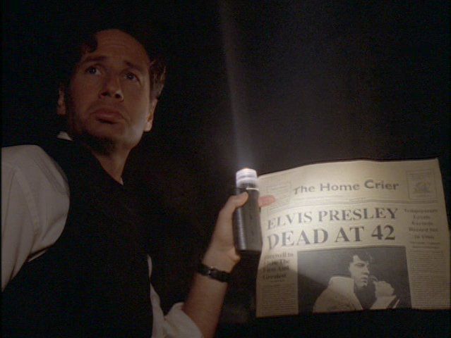 David Duchovny as Fox Mulder holding an old newspaper with the headline "Elvis Dead at 42" looking very sad, from the X Files episode "Home"