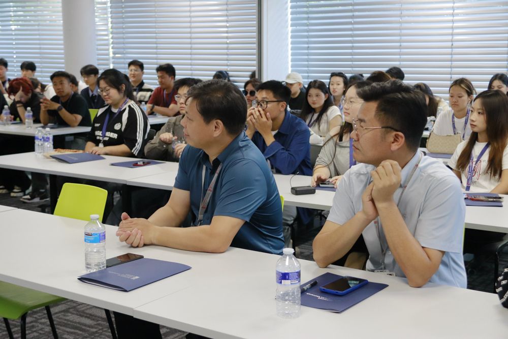 A group of attentive students sitting at desks in a classroom, focusing on a presentation. Some are holding notebooks and bottles of water are visible on the desks.