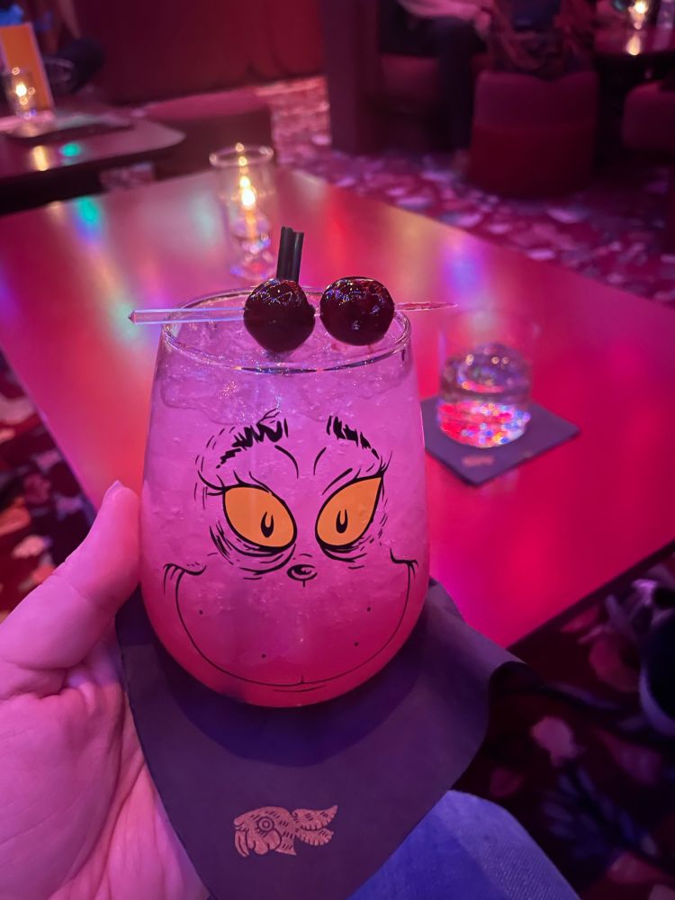 In the foreground, a cocktail in a stemless wine glass with a grinch face on it. In the background, reposado on a single sphere of ice.