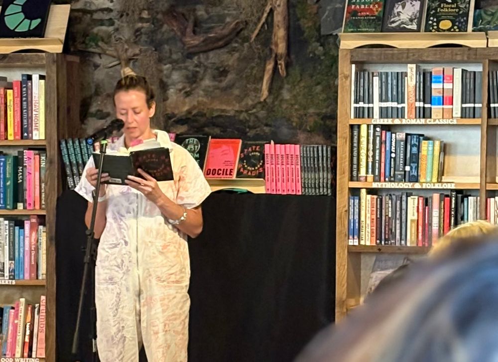 A woman standing between bookshelves at a microphone reading from her novel 