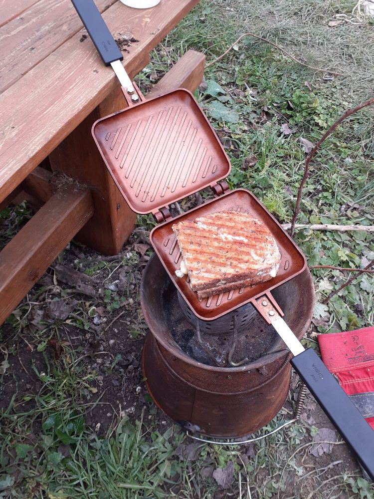 Toasted sandwich being made outdoors over a homemade meths stove in a rusty bucket. 