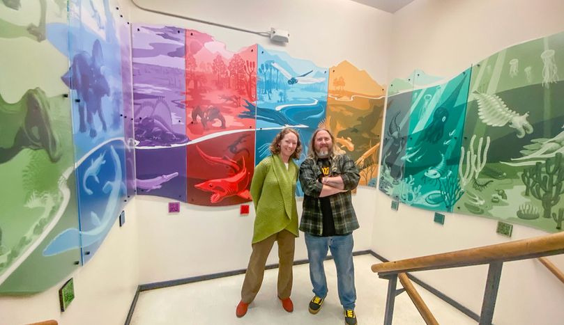 Two people standing and smiling in a corner stairwell. The wall of the stairwell is covered in a colorful mural that weaves around the wall and shows different species of prehistoric lifeforms.