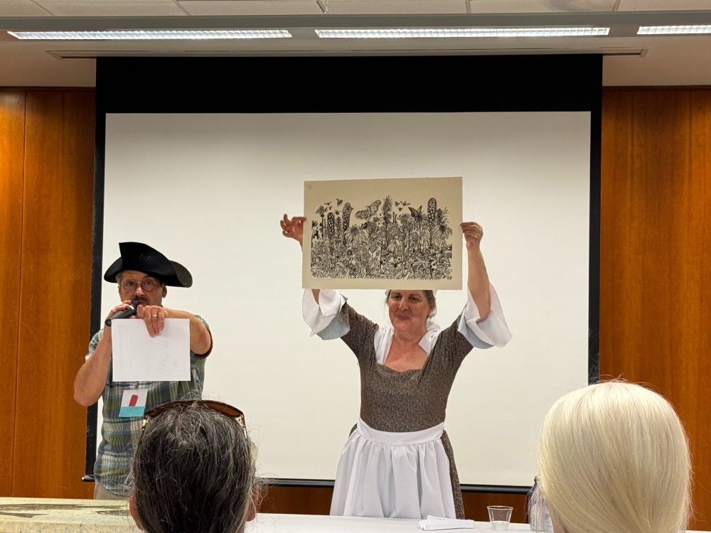 A photo of a man wearing a tricorn hat holding a microphone and pointing at the audience accompanied by a woman dressed in a period costume holding a handmade print by Carrie Carlson above her head