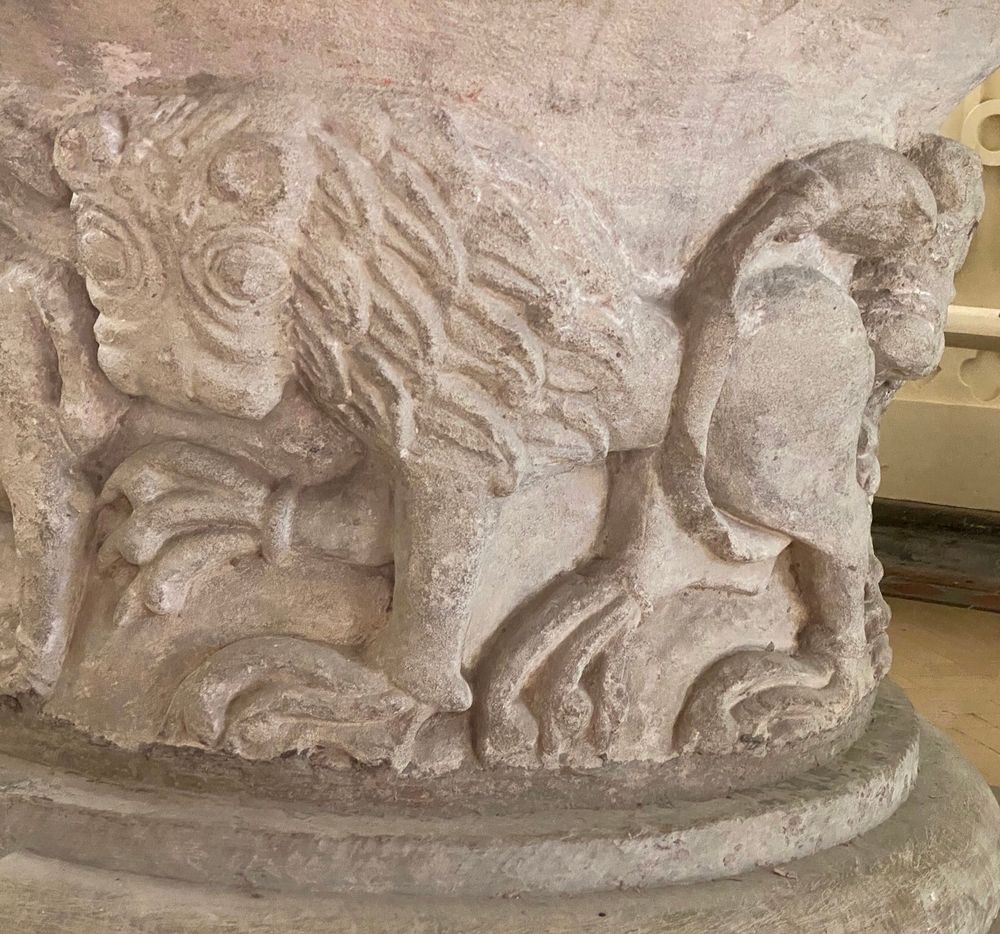 Stylised lion carved in deep relief on the curved side of a pale buff stone font 