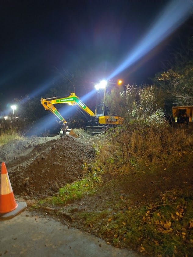 A small yellow JCB digger at the top of a small embankment above a cycle/walking path, digging in the dark under floodlights.