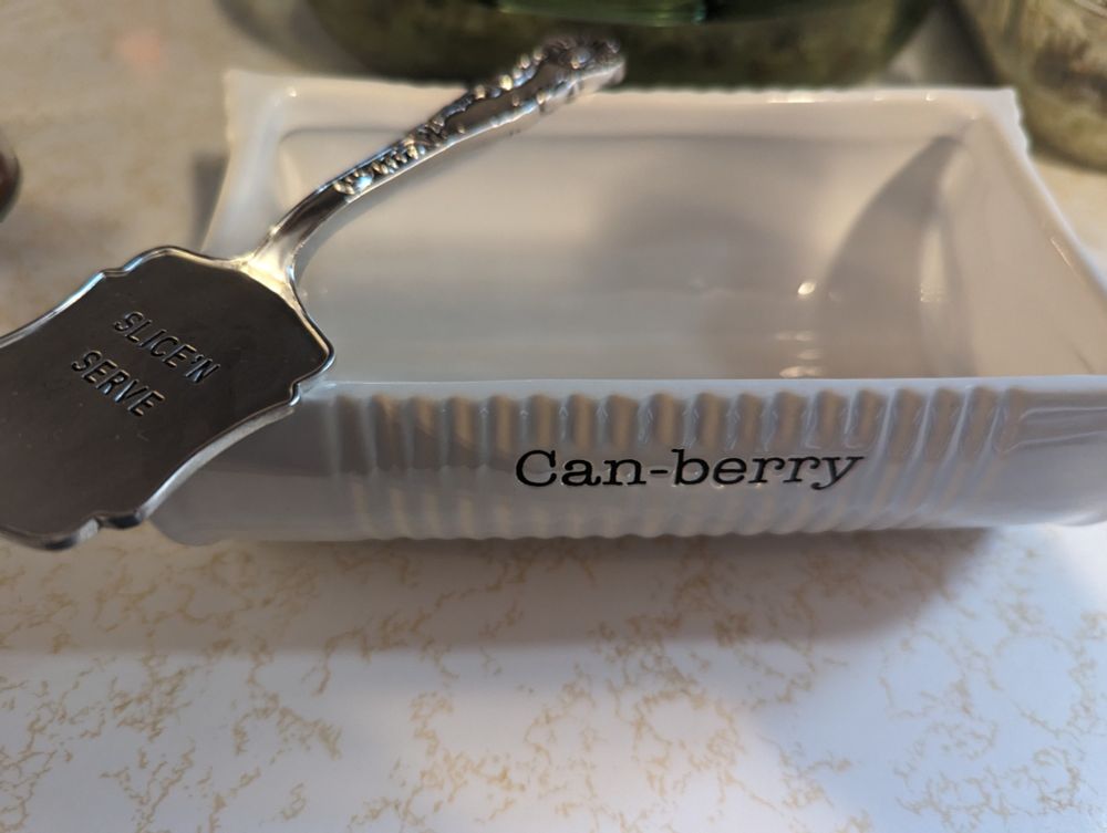 A white porcelain dish shaped like half of a tin can, sliced lengthwise; printed on the front in typewriter-like font is the word "Can-berry". Resting on top of the dish is a silver, spatula-shaped utensil inscribed with the words "Slice 'N Serve" in all capital letters. 