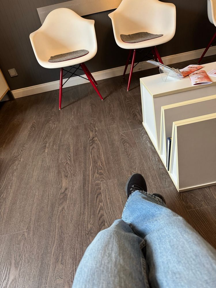 Dunkler Holzfußboden , weiße Stühle und ein Podest, im Vordergrund sind meine Hose und Schuhe zu sehen 