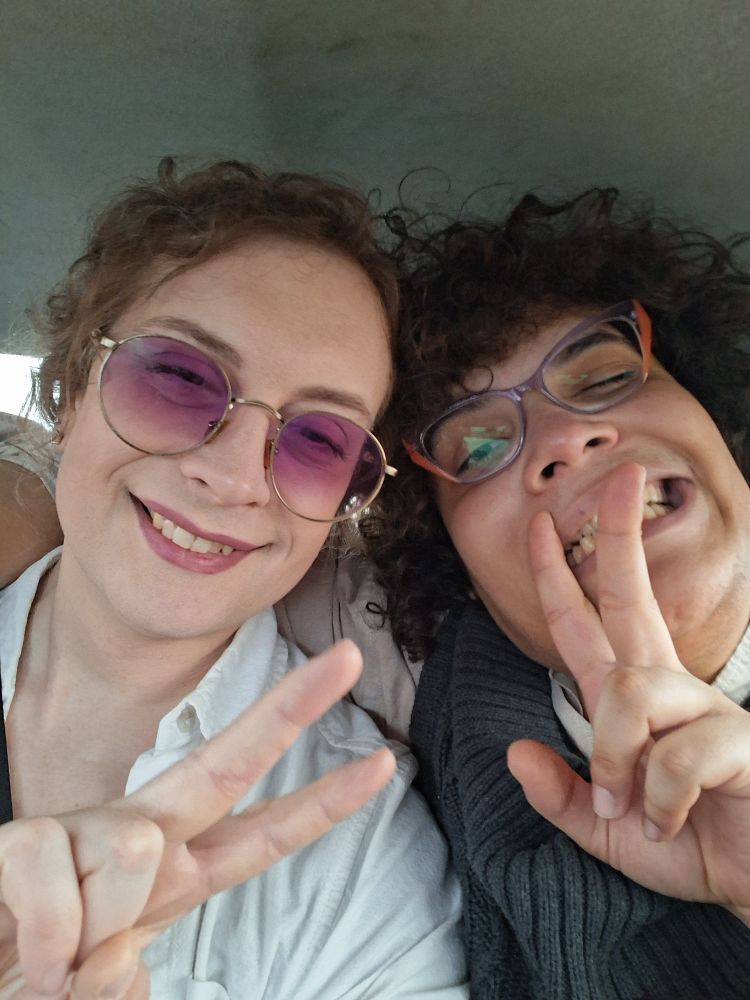 two trans girlies smiling and flashing v for victory signs.