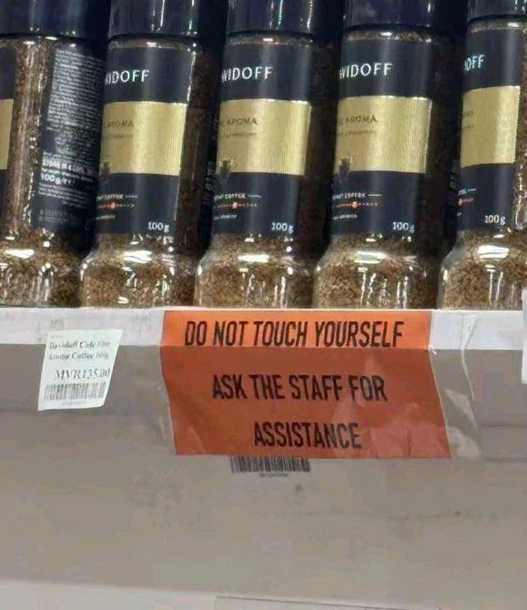 A sign in a store on a white shelf with black text on an orange background reads "Do not touch yourself / Ask the staff for assistance." The products appear to be in black labeled spice jar shaped containers. 