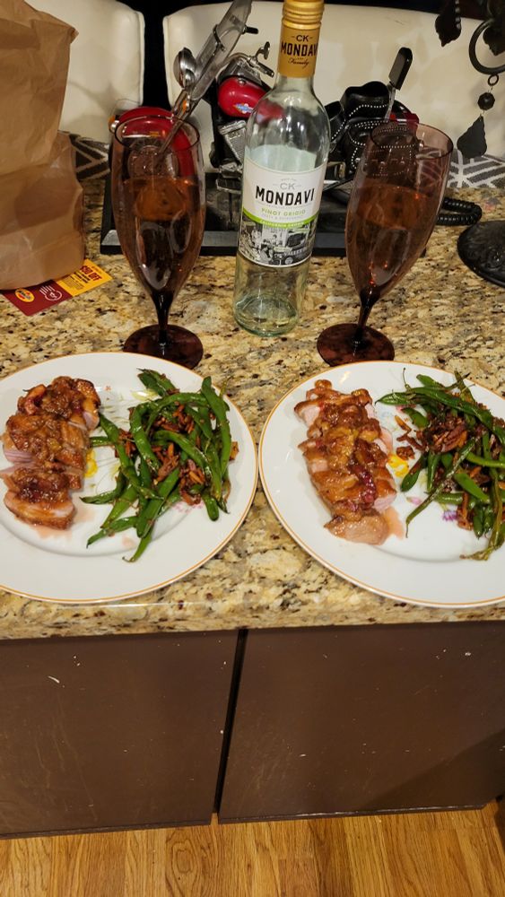 A marble top kitchen counter. On it, two white plates with servings of duck sliced and sauced and the haricot vert almondine. Two violet wine glasses 3/4 full and an empty bottle of pinot grigio sit behind them. 