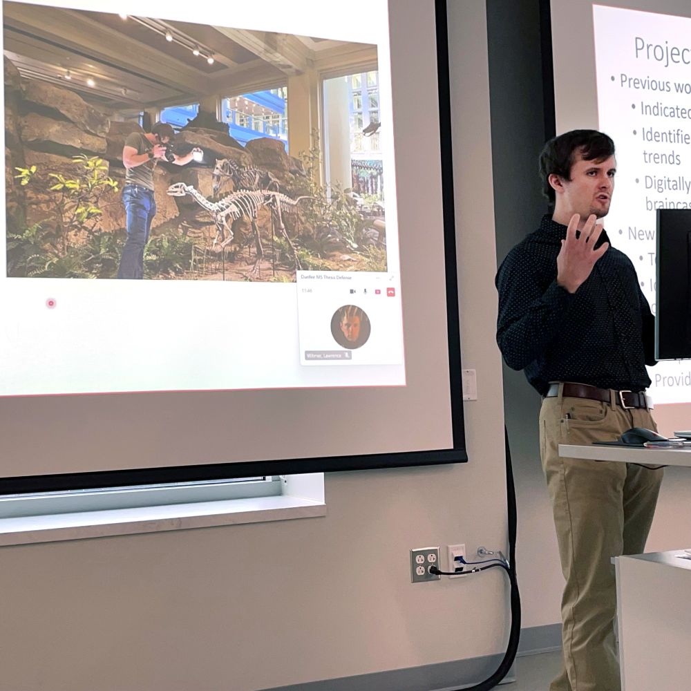 Masters student Daniel Dunfee stands at a podium to give a presentation on his thesis, standing in front of a slide that depicts him in the Carnegie Museum exhibit photographing the Dryosaurus mount.