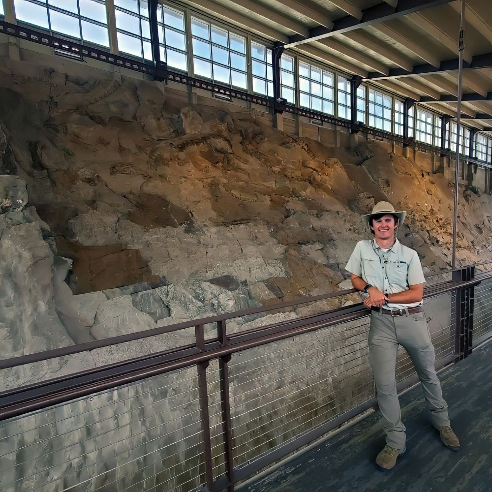 Masters student Daniel Dunfee posing in front of the wall of in situ dinosaur bones at the famous Carnegie Quarry at Dinosaur National Monument in Utah.