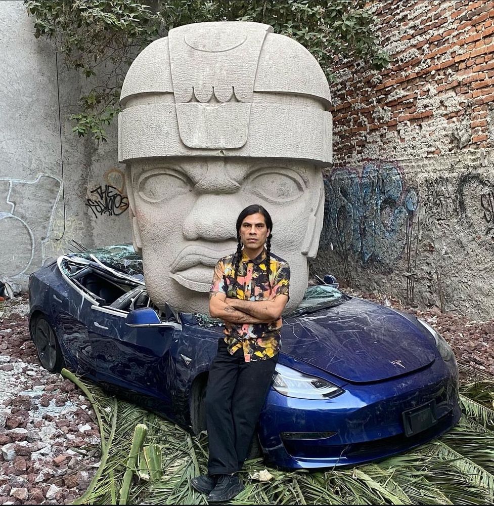 A person leaning against a blue Tesla. The car is completely fucked - roof and windscreen all caved in and flattened - because there is a giant stone head on top of it