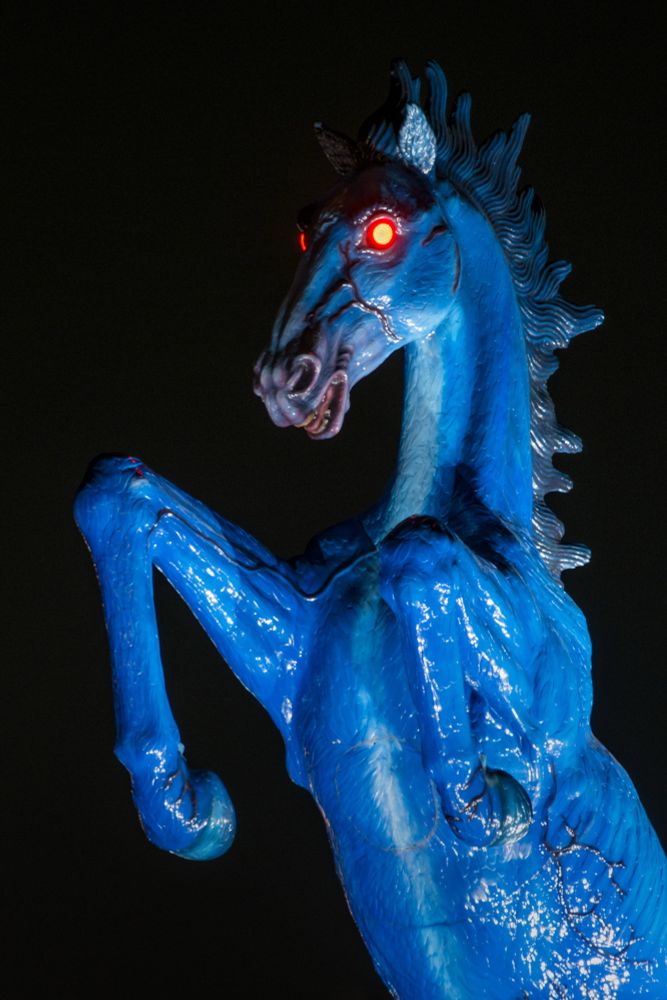 A blue fibreglass horse, reared up on its hind legs, with glowing red eyes. It’s dark so the background is black 