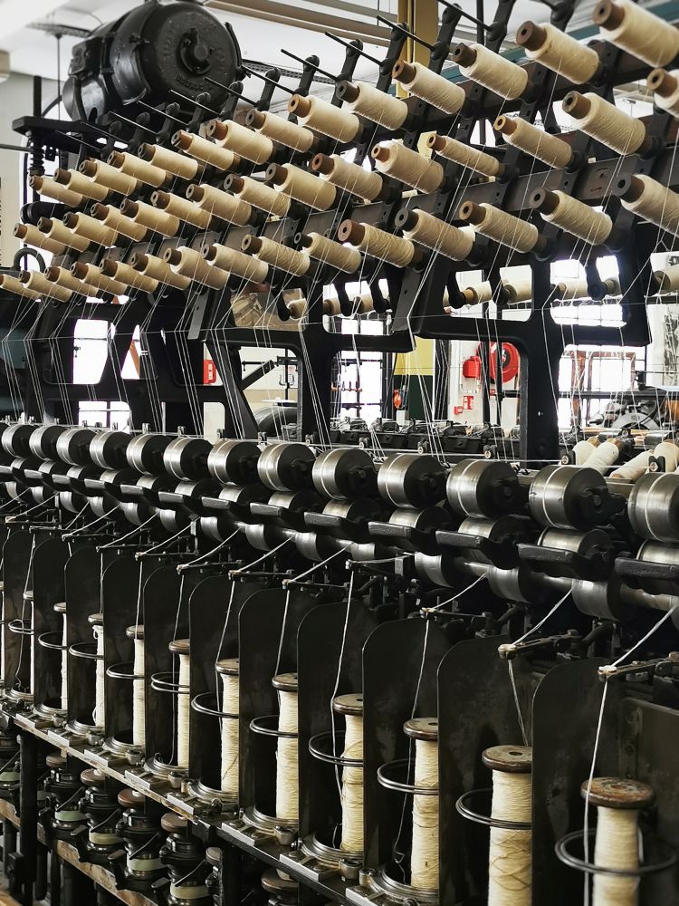 Große alte Maschine aus Metall in einer Spinnerei. Fäden aus hellem Garn führen von den unteren großen Spulen zu den oberen kleineren Spulen. Auf dem Weg dorthin werden sie maschinell verdünnt und verzwirbelt für mehr Festigkeit.