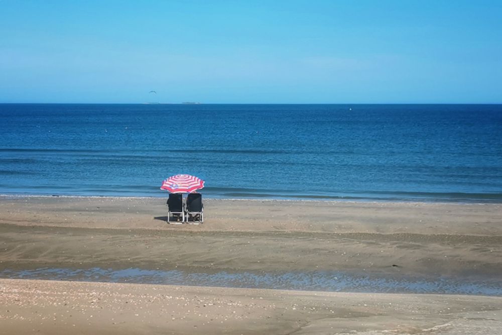 Mit vielen Muschelschalen durchsetzter Sandstrand am hier mal sehr ruhigen Atlantik. Zwei dunkle besetzte Klappstühle und dazwischen ein rot-weiß gestreifter Sonnenschirm stehen einsam im Sand in Blickrichtung Meer. Der Himmel ist blau.